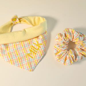 May include: A yellow and plaid dog bandana with the embroidered name "MIX" and a matching scrunchie. The bandana has a yellow collar and a button closure. The scrunchie is made of the same plaid fabric.
