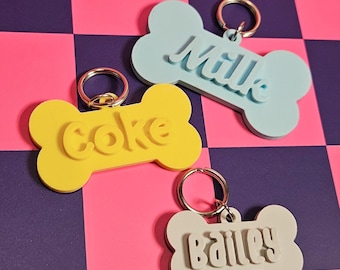 Può includere: Tre targhette per animali domestici a forma di osso in giallo, azzurro e bianco, ciascuna con un portachiavi argentato. L'etichetta gialla dice "Coke", l'etichetta azzurra dice "Milk" e l'etichetta bianca dice "Bailey". Le targhette sono su uno sfondo a scacchi rosa e viola.