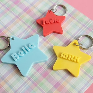 May include: Three star-shaped keychains in red, blue, and yellow. Each keychain has a silver ring and a name or word in a contrasting color: "Sadie", "Bella", and "Milk". The keychains are on a plaid background.