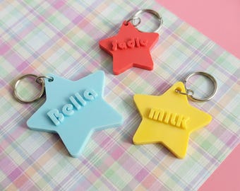 Può includere: Tre portachiavi a forma di stella in rosso, blu e giallo. Ogni portachiavi ha un anello argentato e un nome o una parola in un colore a contrasto: "Sadie", "Bella" e "Milk". I portachiavi sono su uno sfondo a quadri.