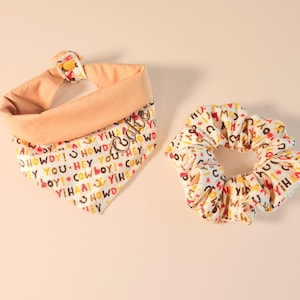 May include: A dog bandana and scrunchie set. The bandana is peach-colored with a white patterned design featuring the words "Howdy," "Hey," "Cowboy," and "Yeehaw." The scrunchie has a matching pattern. The word "Lucky" is on the bandana.