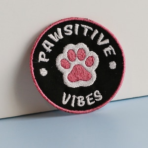 May include: A black and pink embroidered patch with a white paw print in the centre. The text "PAWSITIVE VIBES" is written around the paw print.