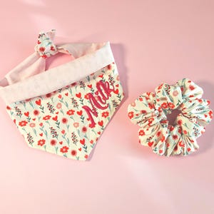 Könnte beinhalten: Ein Set aus Hunde-Bandana und passendem Scrunchie mit floralem Muster. Das cremefarbene Bandana ist mit roten Herzen und Blumen verziert und trägt den Namen "Milk" in Rosa gestickt. Der Scrunchie hat ein ähnliches Blumenmuster. Auf rosa Hintergrund.