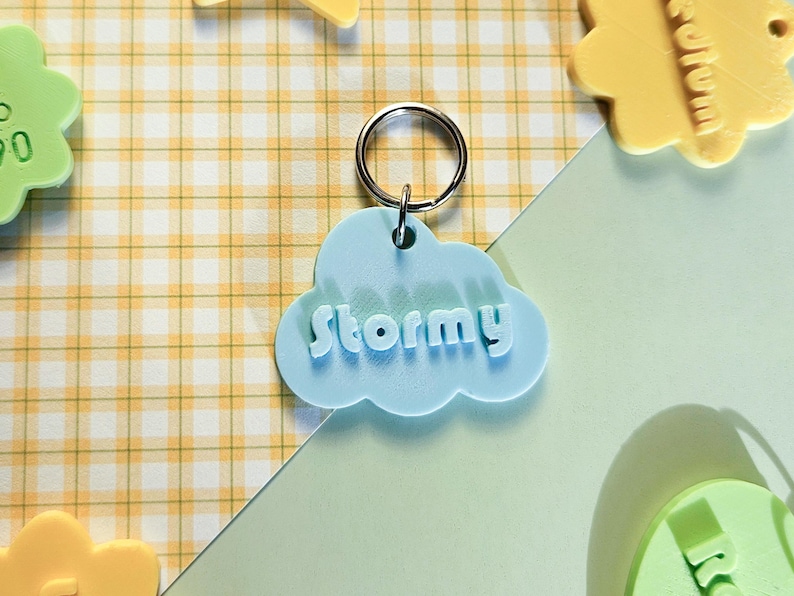 May include: A light blue cloud-shaped dog tag with the name "Stormy" in white, 3D-style lettering. The dog tag has a silver ring and is set against a yellow and white checkered background with other colorful shapes.