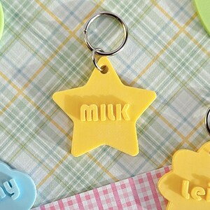 May include: A yellow star-shaped keychain with the word "milk" embossed in the center. The keychain features a silver metal split ring attached to the top point of the star, allowing for easy attachment to keys or other personal items.