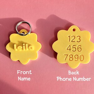 Pu&ograve; includere: Due targhette identificative per animali a forma di fiore giallo. La targhetta a sinistra presenta il nome 'leila' in lettere in rilievo e un anello metallico. La targhetta a destra mostra i numeri '123 456 7890' impressi sulla superficie.