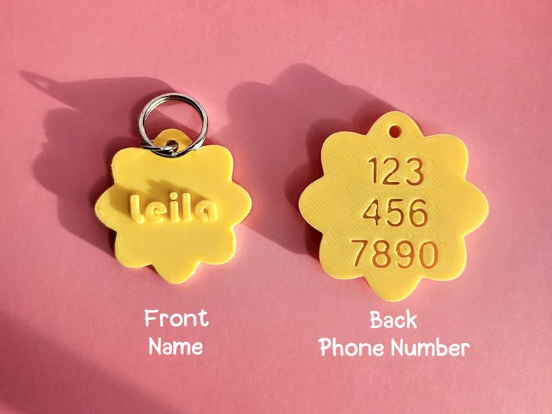 Pu&ograve; includere: Due targhette identificative per animali a forma di fiore giallo. La targhetta a sinistra presenta il nome 'leila' in lettere in rilievo e un anello metallico. La targhetta a destra mostra i numeri '123 456 7890' impressi sulla superficie.