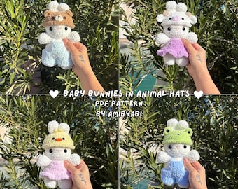 May include: Four crocheted baby bunny dolls, each with a different animal hat: a bear, a cow, a chick, and a frog. The bunnies have white bodies and wear pastel-coloured outfits. The image includes text that reads "Baby Bunnies in Animal Hats PDF Pattern by AmiByAbi."