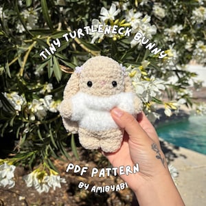 May include: A crocheted bunny with a white turtleneck and small purple accents. The text "Tiny Turtleneck Bunny" is arched above the bunny, and "PDF Pattern by AmiByAbi" is below.