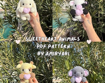 May include: Four crocheted stuffed animals: a bunny, a cow, a giraffe, and an elephant. Each animal is holding a heart. The text "SWEETHEART ANIMALS PDF PATTERN BY AMIBYABI" is displayed in the image.