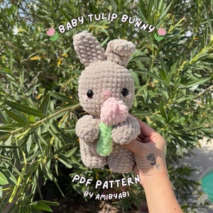 May include: A crocheted bunny holding a pink tulip. The bunny is light brown with black eyes and a pink nose. The text "BABY TULIP BUNNY" and "PDF PATTERN by AMIBYABI" are visible. The background is green foliage.