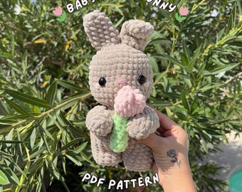 May include: A crocheted bunny holding a pink tulip. The bunny is light brown with black eyes and a pink nose. The text "BABY TULIP BUNNY" and "PDF PATTERN by AMIBYABI" are visible. The background is green foliage.