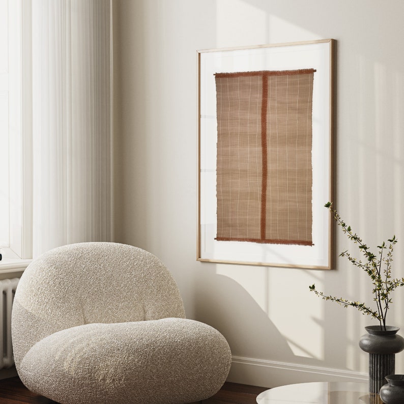 May include: A framed print of a brown woven textile with a red vertical stripe in the center. The print is hanging on a white wall in a room with a white fluffy armchair and a vase with green branches.