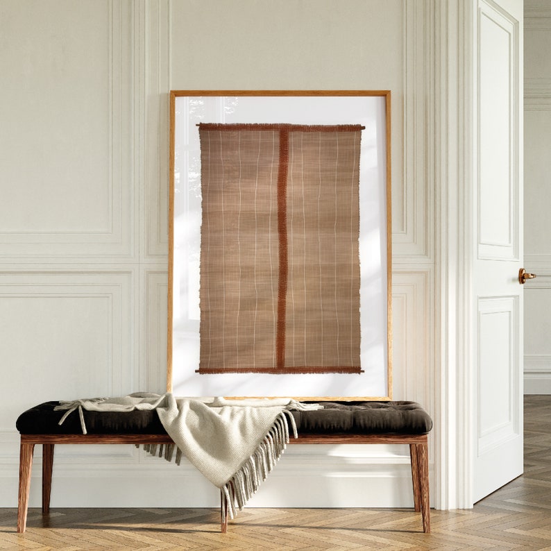 May include: A framed woven wall hanging with a brown and tan color scheme. The hanging is displayed on a white wall above a dark brown bench with a white throw blanket.