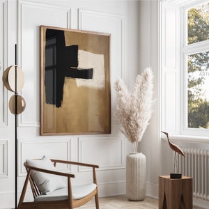 May include: A framed abstract painting with black and white brushstrokes on a gold background. The painting is hanging on a white wall in a room with a chair, a vase of dried pampas grass, and a wooden stool.