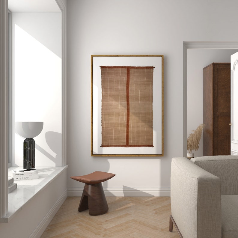 May include: A brown woven wall hanging with a red stripe in the center, framed in a light wood frame, hanging on a white wall in a living room setting.