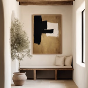 May include: A minimalist abstract painting with black, white, and gold brushstrokes on a canvas. The painting is framed and hanging on a white wall in a room with a wooden bench and a potted plant.