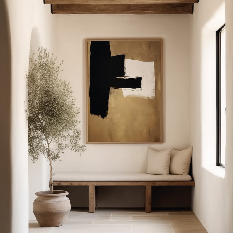 May include: A minimalist abstract painting with black, white, and gold brushstrokes on a canvas. The painting is framed and hanging on a white wall in a room with a wooden bench and a potted plant.