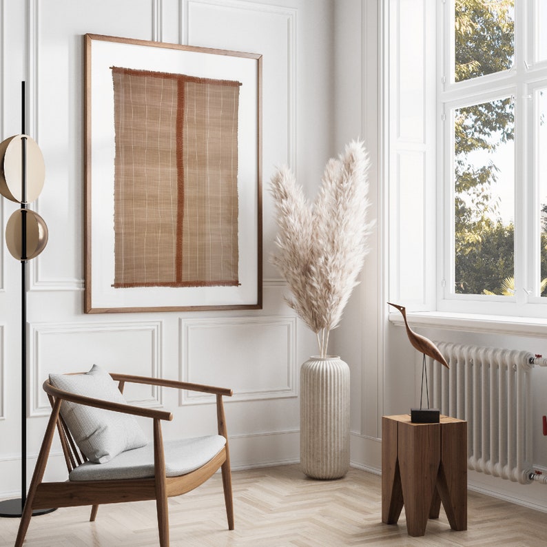 May include: A white room with a wooden chair, a wooden side table, a vase with dried pampas grass, a framed brown and red woven wall hanging, and a wooden bird sculpture.