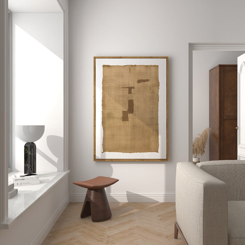 K&ouml;nnte beinhalten: Ein gerahmtes Kunstwerk mit einer braunen gewebten Textur und geometrischen Formen in hellbrauner Farbe. Das Kunstwerk h&auml;ngt an einer wei&szlig;en Wand in einem Raum mit Holzboden und einem beigefarbenen Sofa.