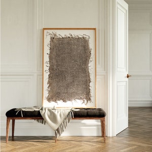 May include: A wooden bench with a brown cushion and a white throw blanket. A framed piece of art with a brown woven texture hangs on the wall behind the bench.