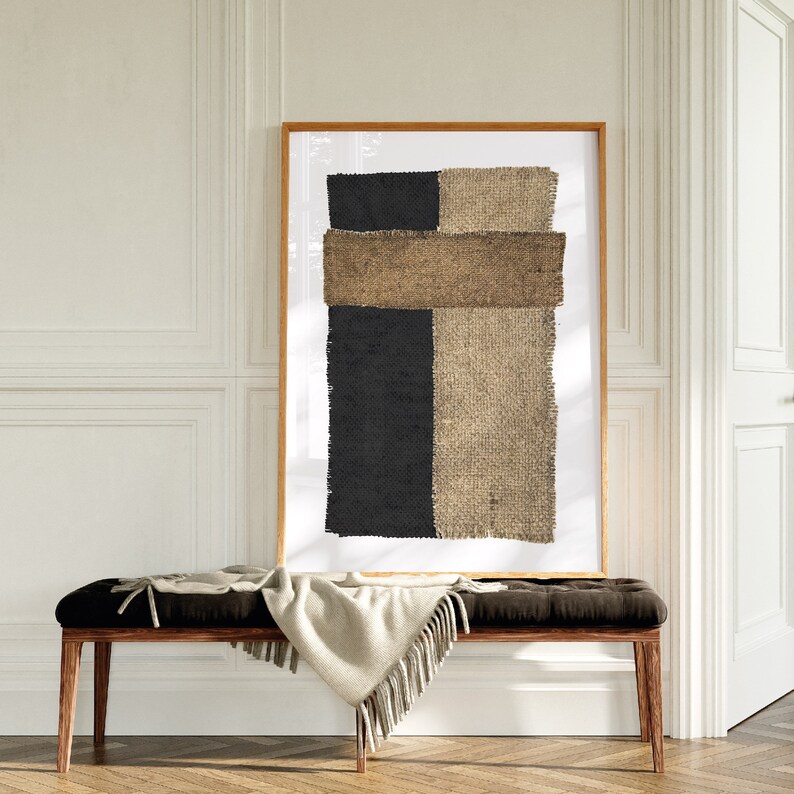 以下が含まれることがあります： 黒と茶色のジュートのデザインが施された額縁付きの抽象的なアートプリント。プリントは白い壁に、白い毛布をかけた木製のベンチの上にかけられています。