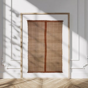 May include: A framed wall hanging with a woven texture in shades of brown and rust. The hanging has a fringe border.