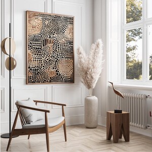 K&ouml;nnte beinhalten: Ein gerahmter abstrakter Kunstdruck mit einem Muster aus schwarzen und braunen Punkten und Linien. Der Druck h&auml;ngt an einer wei&szlig;en Wand in einem modernen Wohnzimmer mit einem wei&szlig;en Stuhl, einer Vase mit getrocknetem Pampasgras und einem Holz-Beistelltisch.