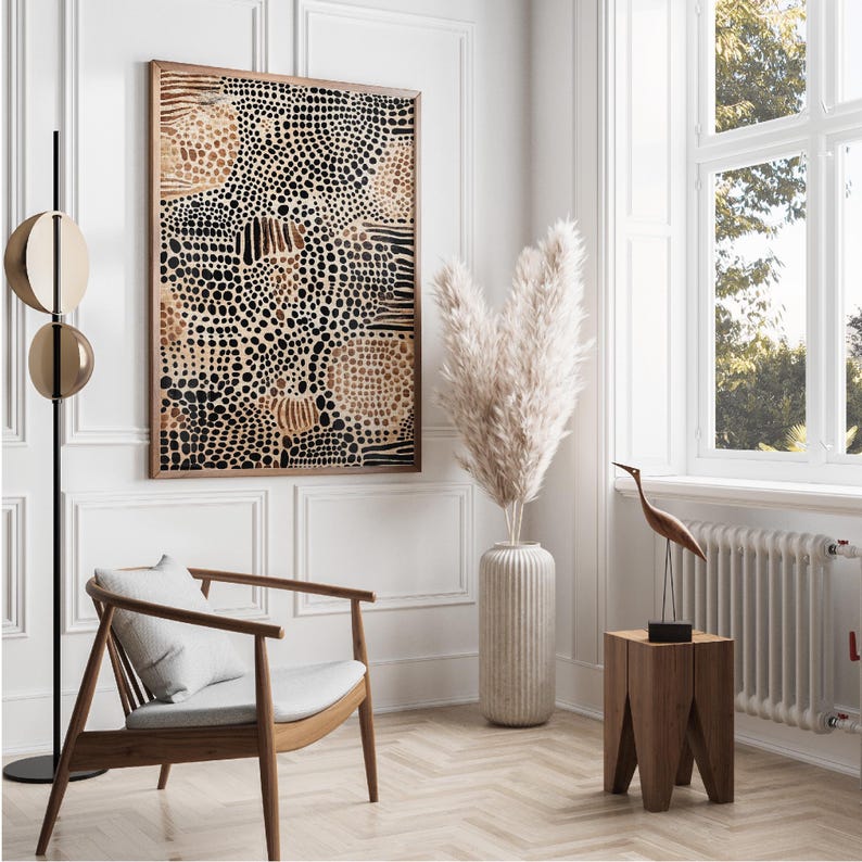 K&ouml;nnte beinhalten: Ein gerahmter abstrakter Kunstdruck mit einem Muster aus schwarzen und braunen Punkten und Linien. Der Druck h&auml;ngt an einer wei&szlig;en Wand in einem modernen Wohnzimmer mit einem wei&szlig;en Stuhl, einer Vase mit getrocknetem Pampasgras und einem Holz-Beistelltisch.