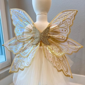 Könnte beinhalten: Weißer Tüll-Tutu mit goldenen Glitzer-Schmetterlingsflügeln. Die Flügel sind schimmernd und haben einen goldenen Glitzer-Umrandung.