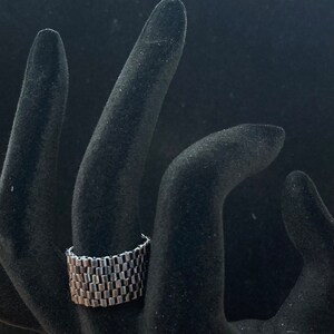 May include: A silver ring with a unique, interlocking chain design. The ring is made of multiple small, rectangular links that are connected together to create a textured, geometric pattern.
