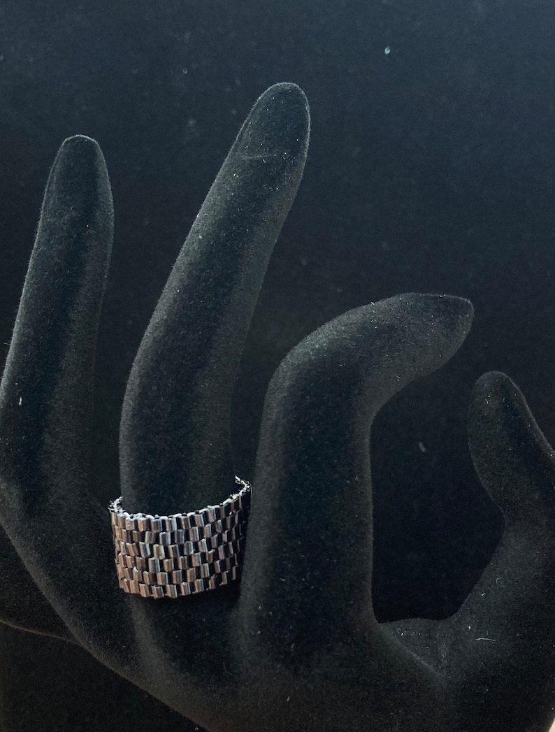 May include: A silver ring with a unique, interlocking chain design. The ring is made of multiple small, rectangular links that are connected together to create a textured, geometric pattern.