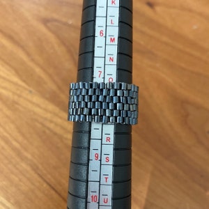 May include: A silver ring with a textured design, measured on a ring sizer that shows sizes K through U.