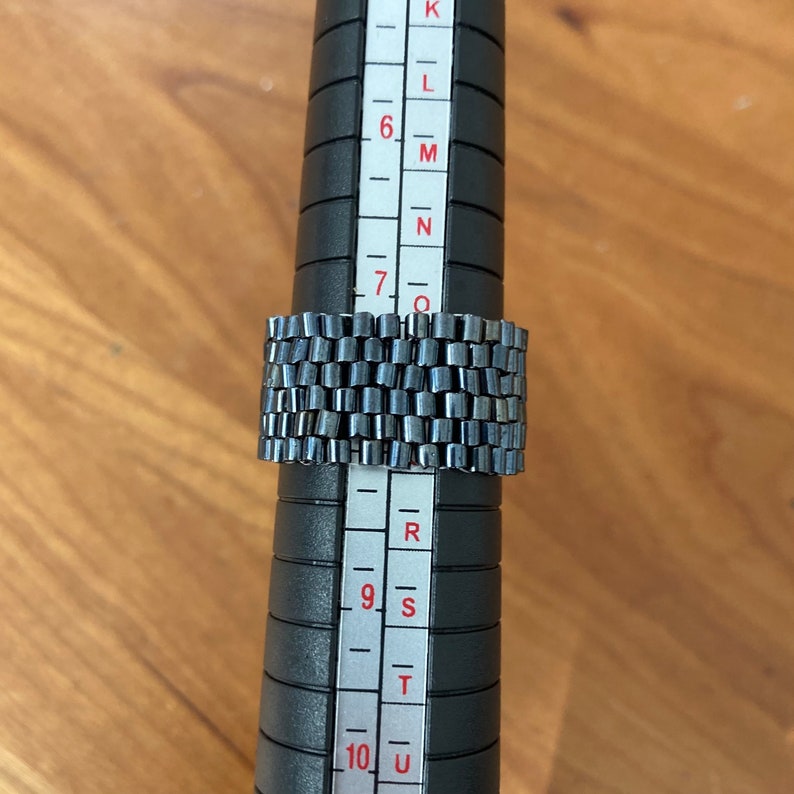 May include: A silver ring with a textured design, measured on a ring sizer that shows sizes K through U.