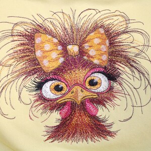 Puede incluir: Un diseño bordado sobre tela de color amarillo pálido que muestra un ave estilizada con plumas marrones alborotadas, ojos grandes con largas pestañas negras, un pico amarillo, carúnculas rosas y un gran lazo naranja con lunares blancos.