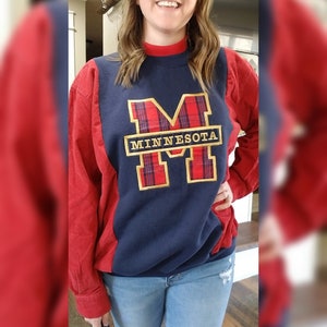 May include: A navy blue and red sweatshirt with the word "MINNESOTA" in gold lettering. The letter "M" is filled with a red plaid pattern. The sweatshirt has red sleeves and a red mock neck.
