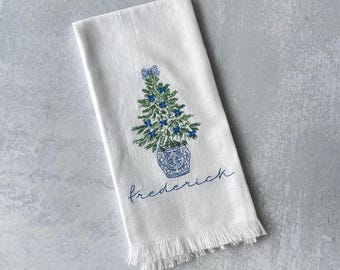 May include: White kitchen towel with a blue and green embroidered Christmas tree in a blue and white pot. The towel has a fringed edge and the text "frederick" is embroidered below the tree.