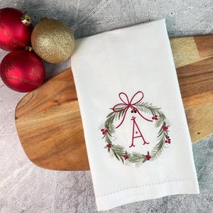 May include: White Christmas-themed hand towel with an embroidered wreath design and the letter "A" in red. The wreath is adorned with green leaves and red berries. Red and gold Christmas ornaments are next to the towel.