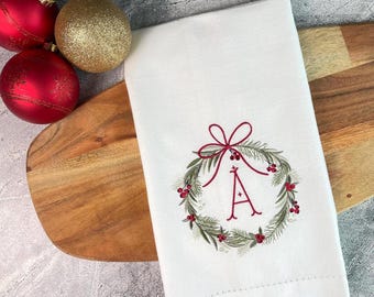 May include: White Christmas-themed hand towel with an embroidered wreath design and the letter "A" in red. The wreath is adorned with green leaves and red berries. Red and gold Christmas ornaments are next to the towel.