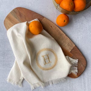 May include: A white kitchen towel with a frayed edge and an embroidered wreath with the letter "H" in the center. The towel is on a wooden cutting board with oranges nearby.