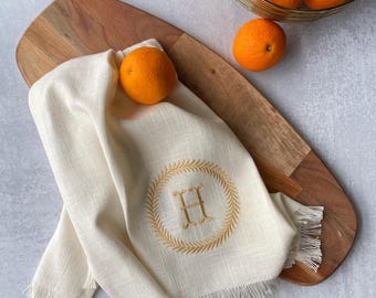 May include: A white kitchen towel with a frayed edge and an embroidered wreath with the letter "H" in the center. The towel is on a wooden cutting board with oranges nearby.