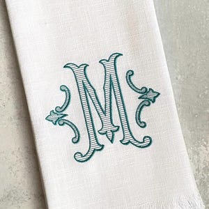 May include: White linen tea towel with a teal embroidered monogram of the letter "M" with decorative flourishes. The towel has a fringe border.