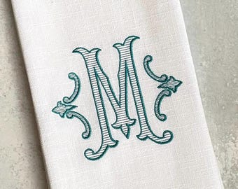 May include: White linen tea towel with a teal embroidered monogram of the letter "M" with decorative flourishes. The towel has a fringe border.