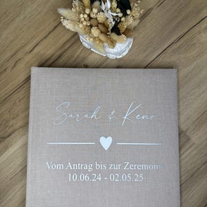 Op de afbeelding: Een vierkant fotoalbum met een beige stoffen kaft, bedrukt met de witte tekst Sarah & Keno, een hartje en de Duitse tekst Vom Antrag bis zur Zeremonie 10.06.24 - 02.05.25. Boven het album staat een wit decoratief voertuig met droogbloemen.
