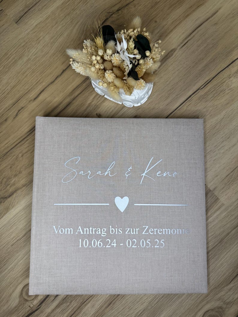 Op de afbeelding: Een vierkant fotoalbum met een beige stoffen kaft, bedrukt met de witte tekst Sarah & Keno, een hartje en de Duitse tekst Vom Antrag bis zur Zeremonie 10.06.24 - 02.05.25. Boven het album staat een wit decoratief voertuig met droogbloemen.