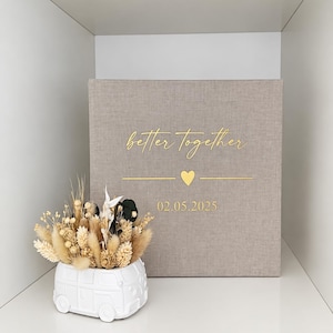 Op de afbeelding: Een vierkant, met beige stof bekleed fotoalbum met goudkleurige tekst "better together" boven een klein hartje en de datum "02.05.2025". Daarnaast staat een witte keramische plantenbak in de vorm van een busje, gevuld met beige en zwarte droogbloemen.