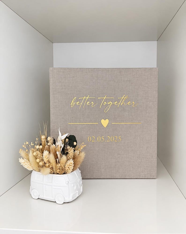 Op de afbeelding: Een vierkant, met beige stof bekleed fotoalbum met goudkleurige tekst "better together" boven een klein hartje en de datum "02.05.2025". Daarnaast staat een witte keramische plantenbak in de vorm van een busje, gevuld met beige en zwarte droogbloemen.