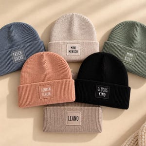 Op de afbeelding: Zes geribbelde gebreide mutsen in blauw, beige, olijfgroen, perzik, zwart en lichtbruin. Elke muts heeft een rechthoekige patch met tekst: Frech Dachs, Mini Mensch, Mini Boss, Sonnen Schein, Glücks Kind en Leano.
