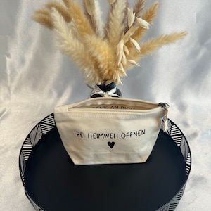 Op de afbeelding: Een beige canvas tasje met een rits en de tekst "Bei Heimweh öffnen" (Duits voor "Open wanneer je heimwee hebt") en een klein hartje.