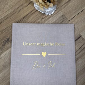 Op de afbeelding: Een vierkant fotoalbum met een lichtgrijze linnen omslag. De voorzijde is voorzien van goudfolie tekst met "Unsere magische Reise" boven een gouden hartpictogram en de tekst "Du & Ich" in een cursief lettertype.